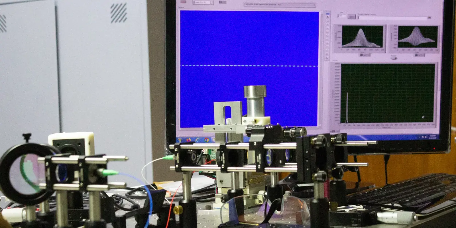 Fotografia de uma mesa de laboratório com um sistema óptico e um monitor.